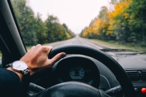 an image of a from the perspective of a person driving who's in a car holding the wheel with his left hand with a silver watch on his wrist on a sunny day. 